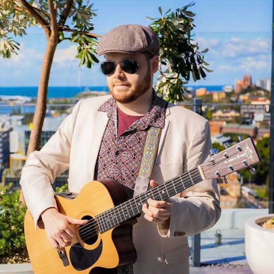 Singer, song writer. Connor Wink A man is standing outdoors holding a guitar. He is wearing sunglasses, a flat cap, and a light-colored jacket over a patterned shirt. Behind him, there's a scenic view of a coastal city under a clear blue sky. There are buildings in the distance and greenery in the foreground.