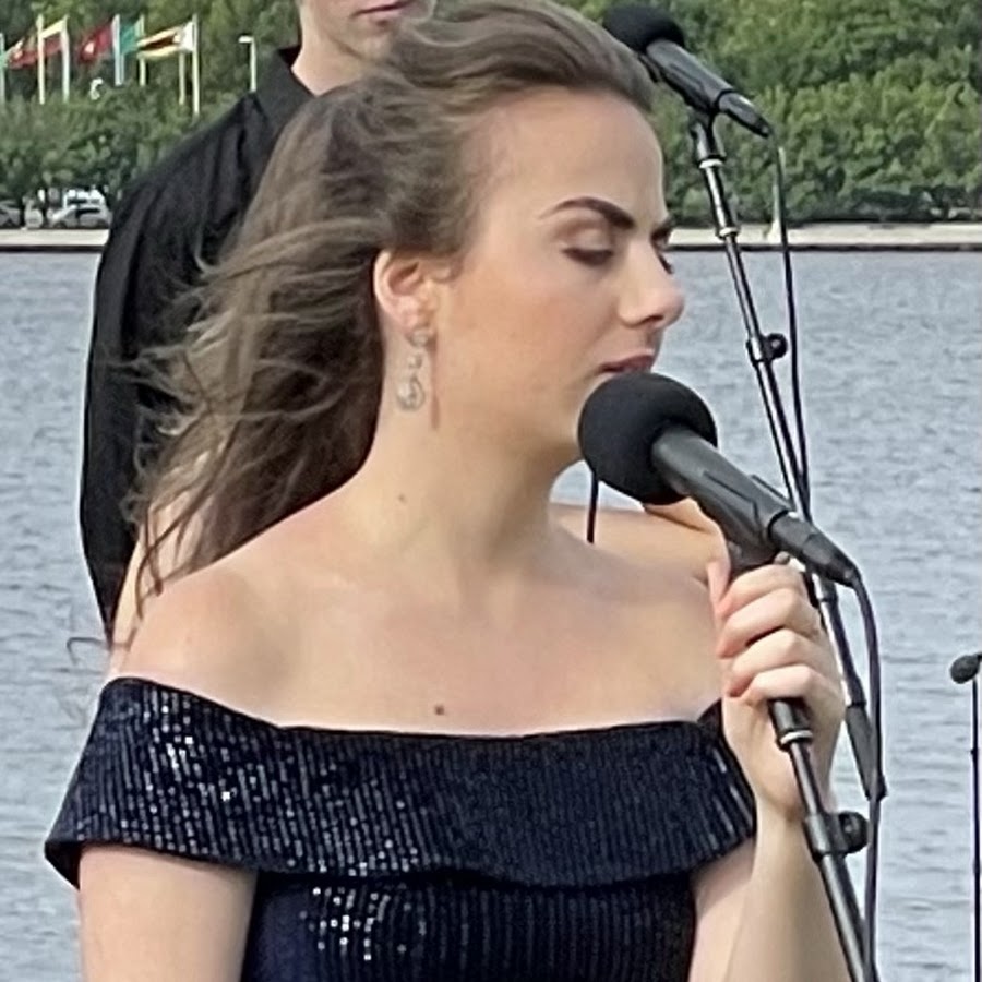 singer , song writer Lara Nakhle The picture shows a woman singing into a microphone. She has long, dark hair that is swept to the side and appears to be wearing makeup. She is wearing a dark, off-the-shoulder top with a glittery texture. In the background, there is a blurred figure of a person and a hint of a natural setting that might be a body of water or greenery. The focus is on the woman, suggesting she is the main subject, likely performing at an event.