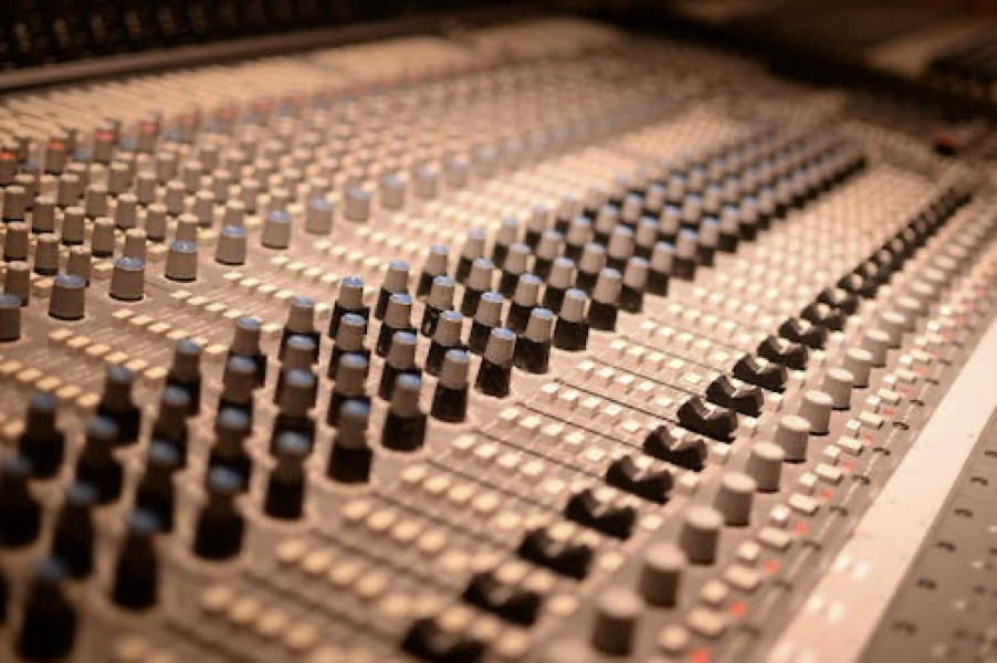The picture shows a close-up of a large audio mixing console. It has numerous vertical sliding controls (faders) and rotary knobs, which are used to adjust the sound levels and audio effects of different input channels. The console appears to be in a studio setting, with a focus on the central part of the board where the faders are densely packed together. The lighting is warm, highlighting the metallic and black colors of the console.