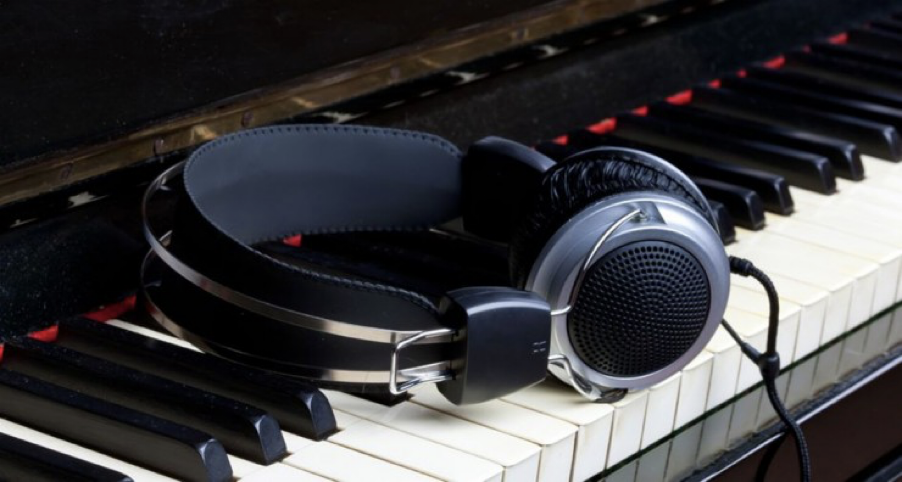 The picture shows a pair of black over-ear headphones resting on the keys of a piano. The piano keys are a mix of white and black, indicating it is a standard keyboard layout. The headphones have a silver metal accent on the earpieces and are connected by a black headband. There is a cable attached to the headphones that drapes over the keys. The piano keys reflect a bit of light, suggesting a glossy finish.